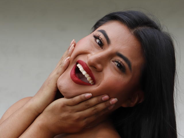 MichelleDenise with dark hair, red lipstick, and a smile, holding her face with hands, showing off her teeth, and looking cute with a sexy, happy expression.