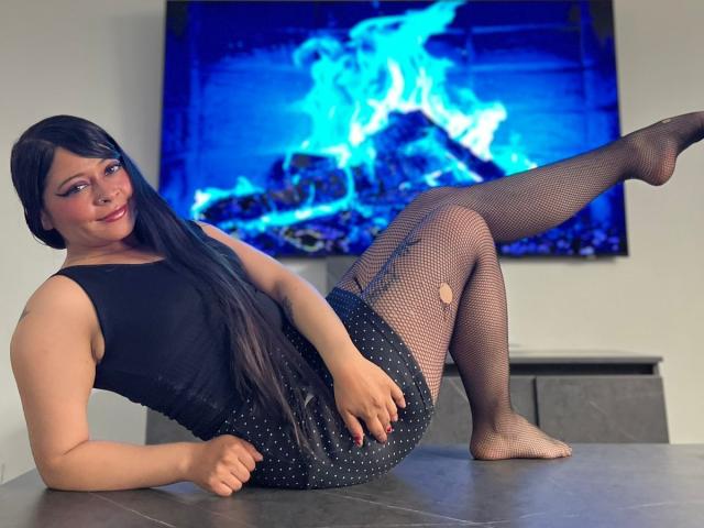 SophiEswett with long black hair, black sleeveless top, black polka dot skirt, fishnet stockings, and a butt hole in one leg, laying on a table, leg raised.