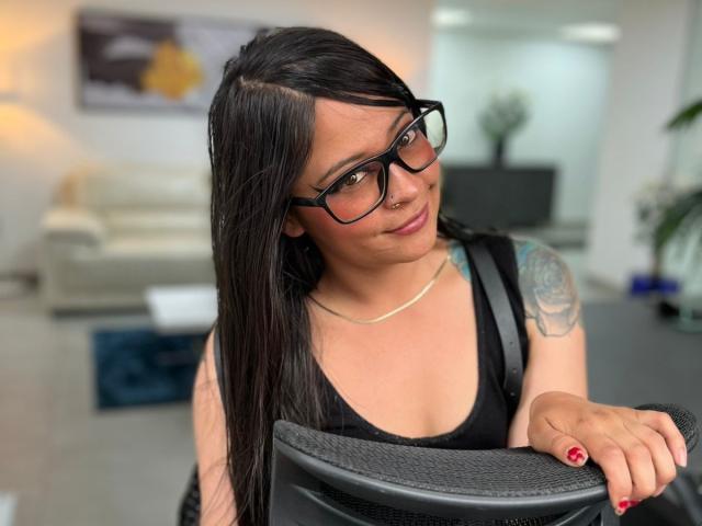 SophiEswett with long black hair, glasses, nose ring, black tank top, tattoo on left arm, red nail polish, leaning on black chair, smiling slightly, looking at camera.