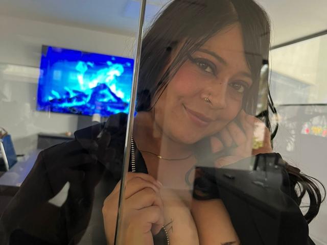 SophiEswett with black hair, nose ring, and subtle makeup, smiling at the camera, holding her phone, wearing an open black shirt, showing her big boobs, through a glass reflection.