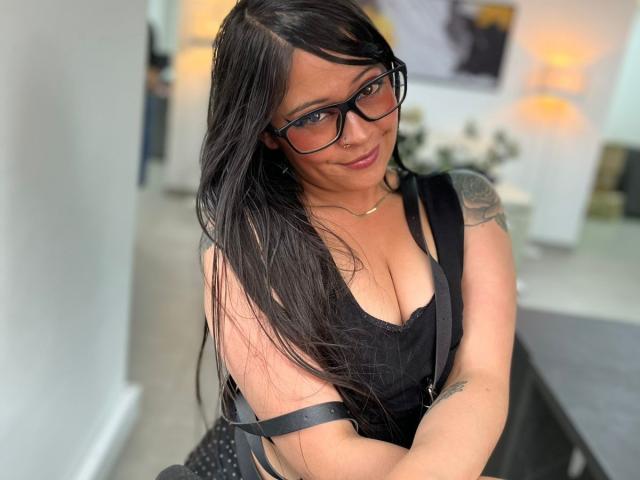 SophiEswett with long black hair, glasses, black tank top showing her tits, tattoo on right arm, smiling, holding black straps, standing in a modern room with lamps.