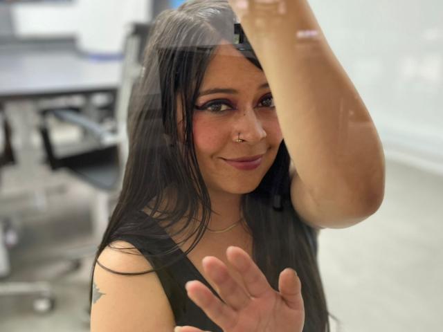 SophiEswett with long black hair, purple eyeshadow, nose ring, smiling, wearing a black sleeveless top, raising her right arm, showing off her tits, in a bright room.