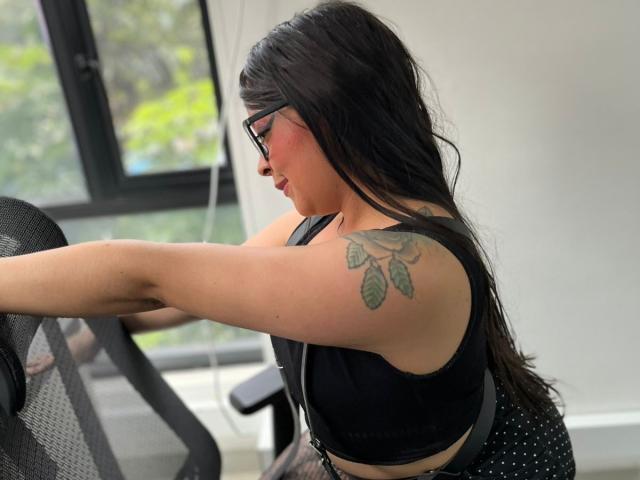 SophiEswett with black hair, glasses, black bra, and polka dot shorts, tattoo on upper arm, leaning on black chair, window in background.