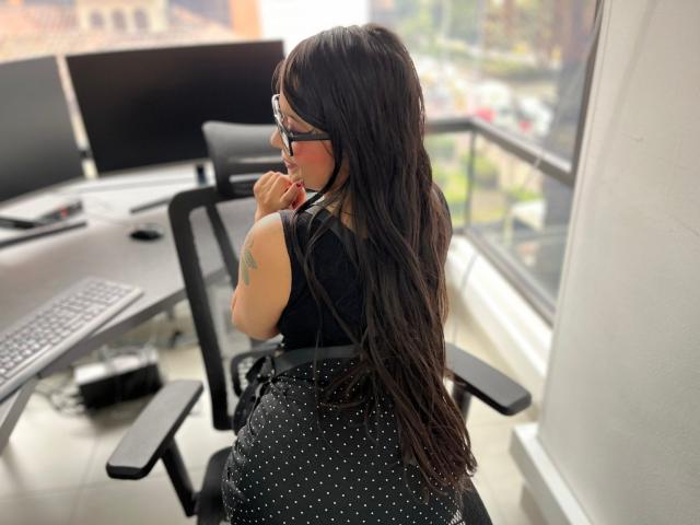 SophiEswett with long dark hair, wearing glasses, black sleeveless top, and polka-dot skirt, sits in office chair, hand near mouth, showing a tattoo on her right arm.