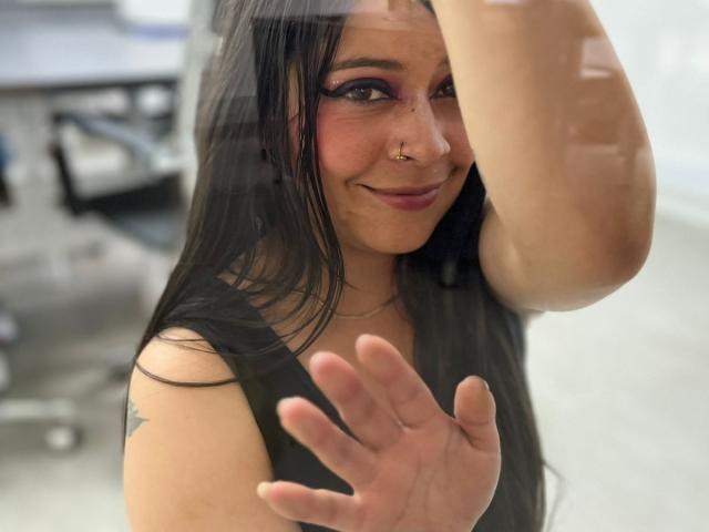 SophiEswett with long black hair, nose ring, and purple eyeshadow, smiling at the camera, hand raised, wearing a black top, visible tattoo on her left shoulder.