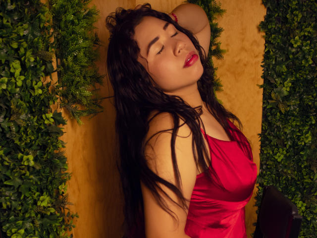 Susie Willianson, con lunghi capelli neri, occhi chiusi, indossa un abito di seta rossa, appoggiata a un fogliame verde, braccia sollevate sopra la testa, labbra leggermente dischiuse, con un'aria rilassata e sensuale..