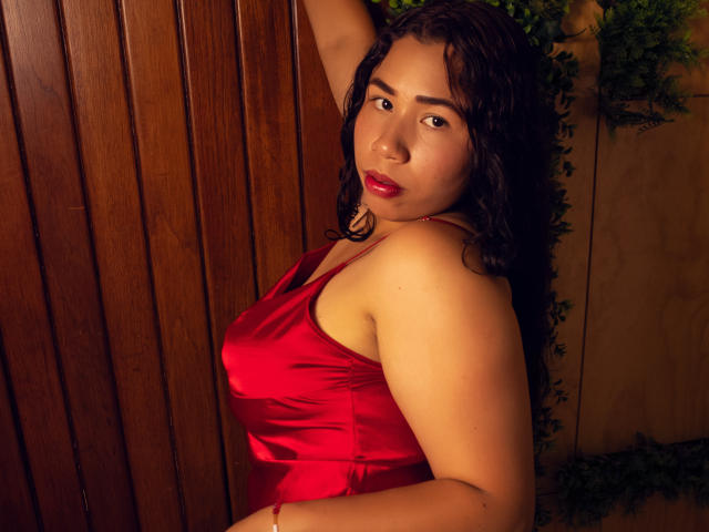 SusieWillianson in a red satin dress, arms raised, wavy black hair, red lipstick, looking at camera, wooden wall, green plant, tan skin, curvy, soft lighting, sexy.