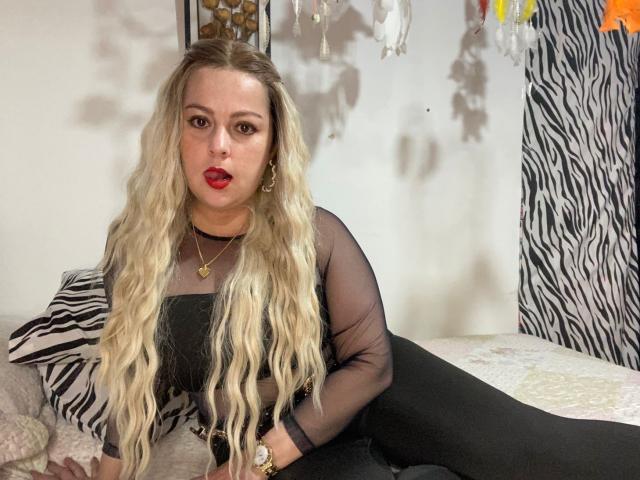 SamanthaGold with long wavy blonde hair, red lipstick, black sheer top, black pants, sitting on bed, zebra-patterned pillow, gold necklace, gold earrings, looking at camera.
