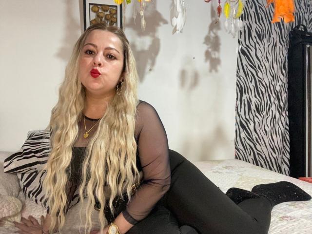 SamanthaGold with long blonde wavy hair, wearing a sheer black top and black pants, pouting her red lips, lying on a bed with zebra-patterned pillows, wearing a gold necklace.