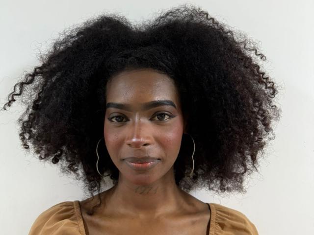 AriannyOrtega has a big, fluffy, curly black afro, dark brown skin, and is wearing a tan top with hoop earrings, looking straight at the camera with a slight smile.