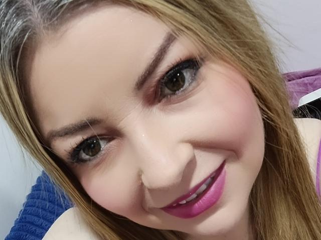 NatalyHami a les cheveux blonds, les yeux marrons et porte un rouge à lèvres rose. Elle sourit, la tête légèrement inclinée sur le côté, dévoilant son joli visage et son maquillage discret..