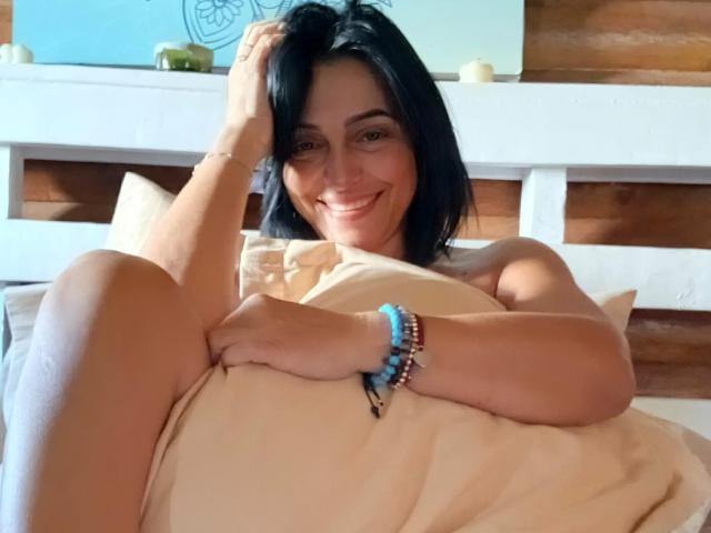 ZullyXHot with black hair, smiling, lying in bed, holding a pillow, showing her ass, wearing a blue bracelet, and a bracelet with beads on her right wrist.