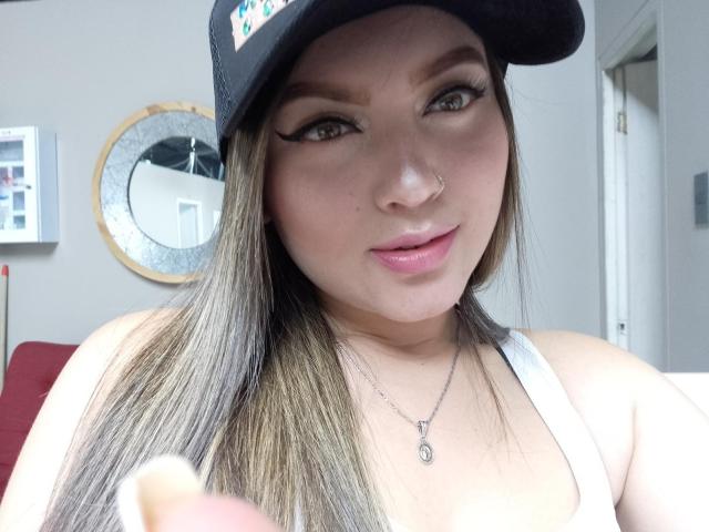 LillithDeluxe with straight brown hair, black cap, white tank top, nose ring, and winged eyeliner, holding a pink object, smiling slightly, light skin, green eyes, pink lips.