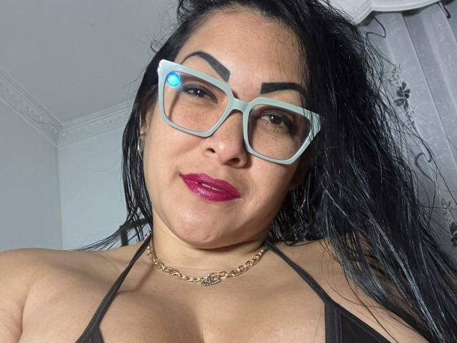 Mellissa Cortez mit dunklem Haar, pinkem Lippenstift, dicken Augenbrauen, blauen Brillen, einer Goldkette, einem schwarzen Top mit Halter, das ihre Brüste zeigt, leicht zerzaustem Haar, lächelnd, im Innenbereich..