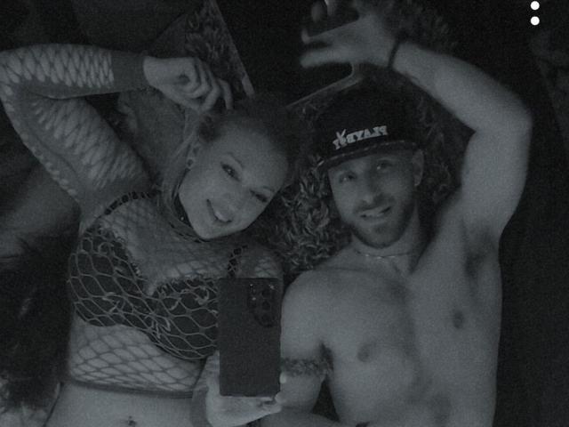 Black-and-white selfie of CloudyCouple, a blonde woman with large boobs in fishnet top and a shirtless man with a hat, lying down and smiling at the camera.