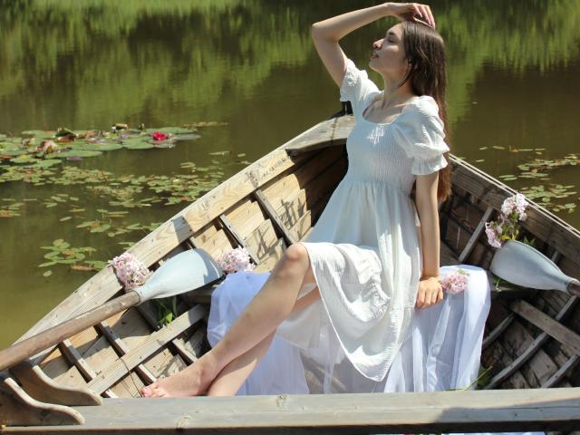 Bluelia, vestita di bianco, seduta su una barca di legno, con un braccio alzato che mostra i suoi seni, gambe incrociate, fiori sulla barca, acqua con ninfee sullo sfondo, una giornata di sole..