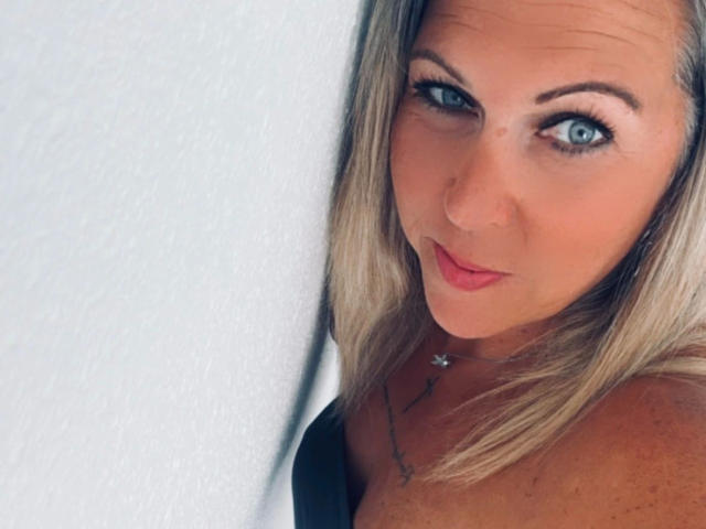 AurelieNoir-hot with blonde hair, blue eyes, and pink lipstick, showing off her bare shoulders and black strap, wearing a delicate necklace. Her tits are slightly visible.