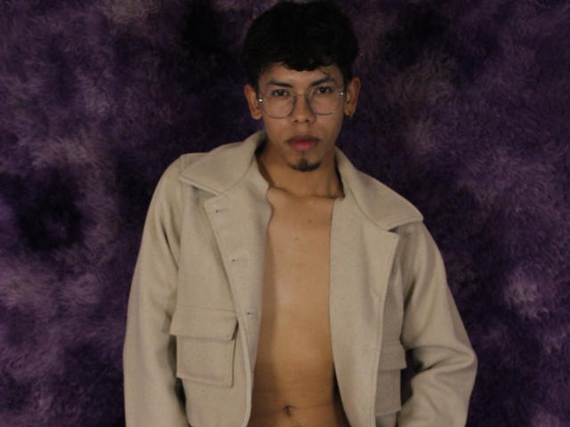 LianStonne with glasses, short black hair, light brown skin, small goatee, shirtless, beige coat, purple fuzzy background, looking straight at camera, hands by sides.