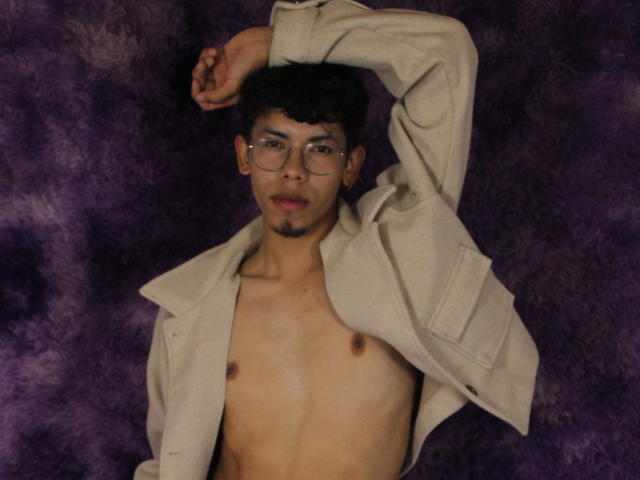 LianStonne, topless, with glasses, and a beige jacket, has a slight goatee and a relaxed, confident expression, lying down against a purple, fuzzy background.