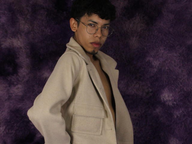 LianStonne with glasses, short curly black hair, light brown skin, red lips, wearing a beige jacket, chest exposed, purple textured background, standing with hands on hips.