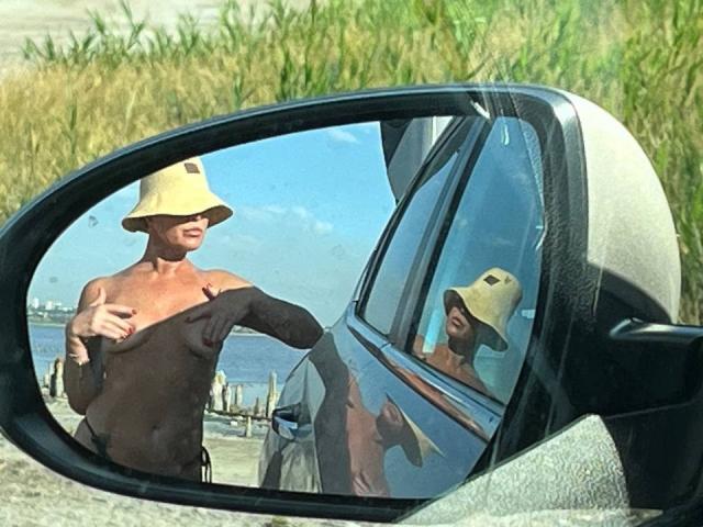 PleasureWeCouple's reflection in the car mirror shows a shirtless, tan-skinned dude with a yellow bucket hat, pulling down his black shirt, revealing his chest.