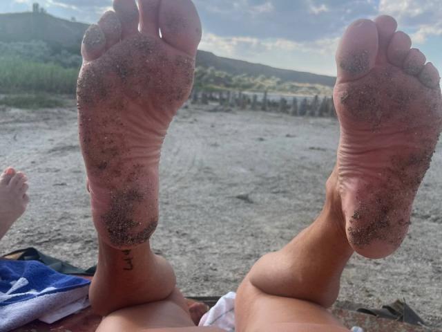 PleasureWeCouple's dirty soles facing the camera, sand stuck to their toes, with a small black tattoo on the left ankle. Feet are up, showing a relaxed beach vibe.