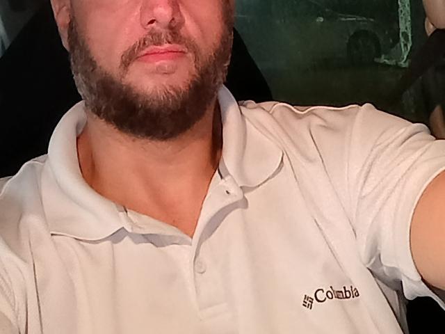 DirtyLegforYou with a beard, wearing a white polo shirt, has a slightly flushed face, and is taking a selfie. His shirt has a small logo on the left.