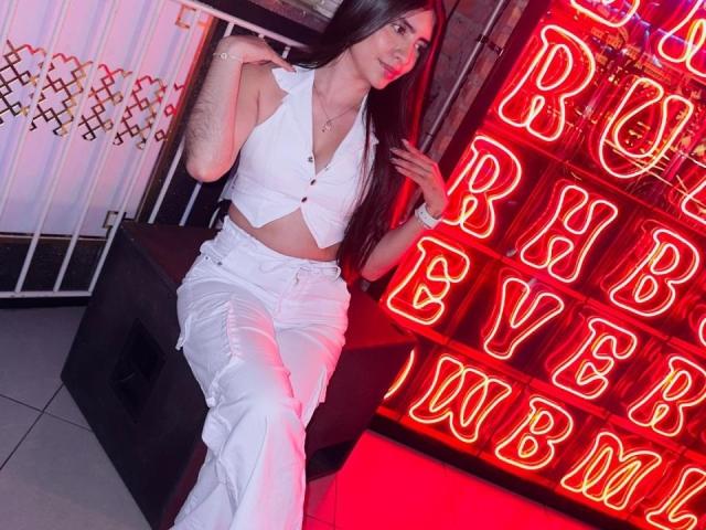 LuanaJoys with long dark hair, white sleeveless crop top, and white pants, sitting on black bench, neon red letters in background, right arm raised, left arm resting.