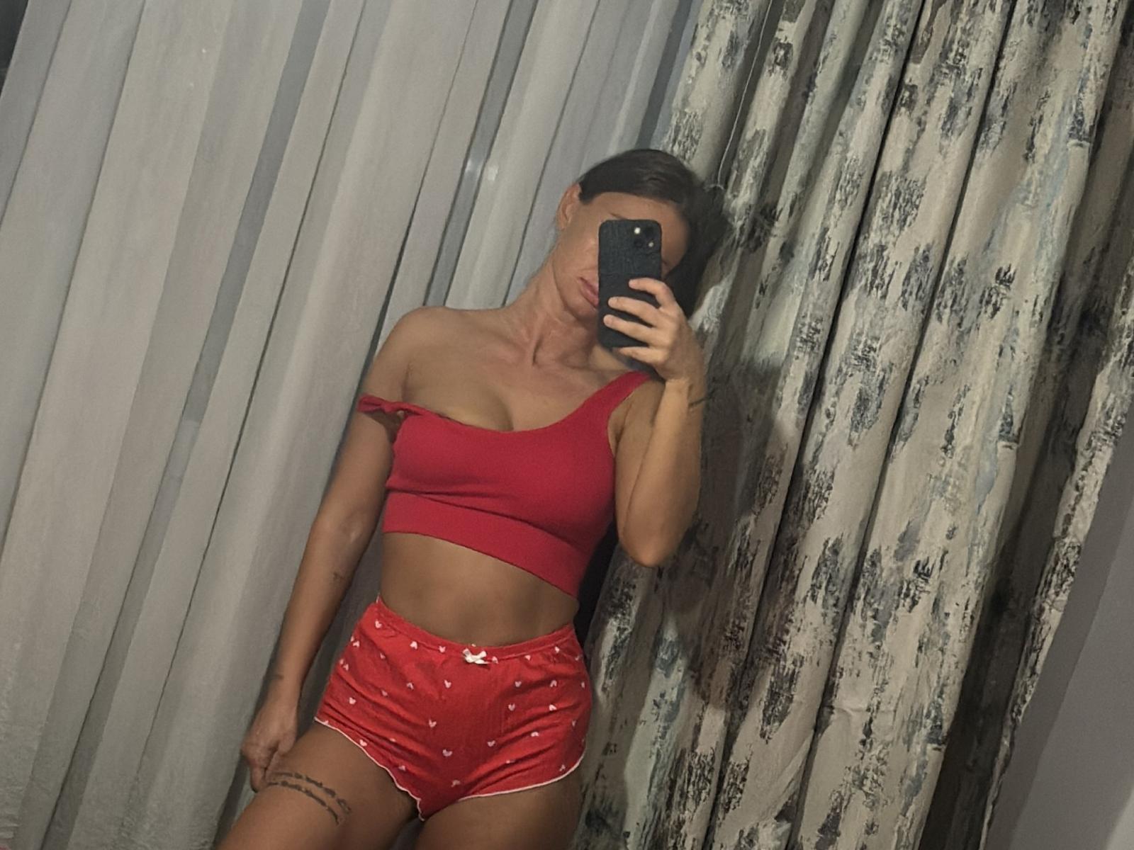 JesssicaJesss with small tits, wearing a red one-shoulder crop top and shiny red panties, taking a mirror selfie, holding a black phone, in a dimly lit room.