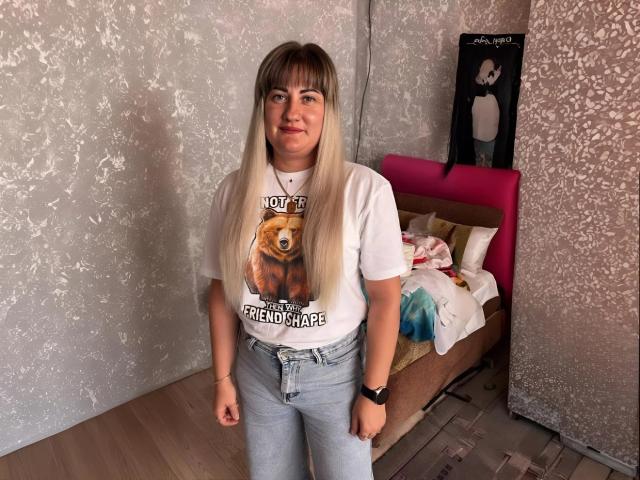 LucyWow with long blonde hair, white t-shirt with bear graphic, blue jeans, standing in room, pink bed, white wall, wooden floor, black wristwatch, casual, slightly chubby, neutral expression.