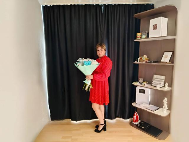 LucyWow in a red dress, holding a bouquet of blue and white flowers, standing in a room with black curtains, wearing black high heels, short blonde hair.