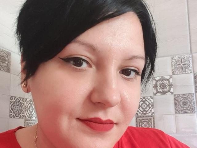 MayaBlowsYou, avec ses cheveux noirs, son rouge à lèvres rouge et son eye-liner ailé, porte une chemise rouge, a un visage légèrement rougi et mignon, et regarde l'appareil photo..