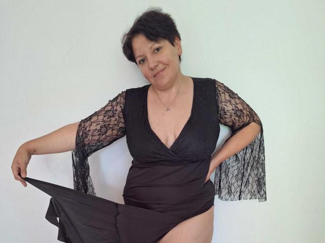 MayaBlowsYou, une femme aux cheveux noirs courts, portant une robe noire en dentelle avec un décolleté en V plongeant, tirant la jupe vers le bas pour révéler ses sous-vêtements noirs, souriant à la caméra..