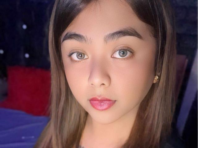 Una foto di FuckMeSoHard69 con la pelle marrone chiaro, gli occhi verdi, i capelli castani lisci, il rossetto rosa e un trucco leggero, che guarda direttamente l'obiettivo con un'espressione neutra..