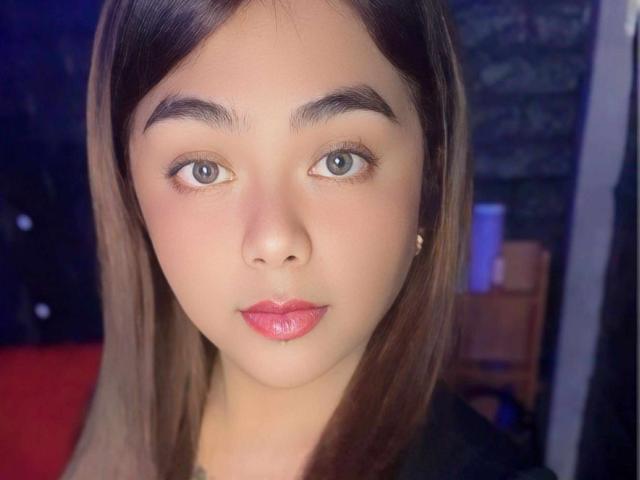 FuckMeSoHard69 tiene una cara bonita con piel marrón claro, cabello castaño lacio, ojos verdes y labios rosados y brillantes. Lleva una camisa negra y tiene pequeños aretes..
