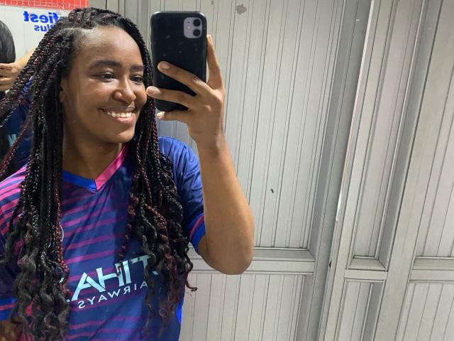 NaomiReyes with long, dark braided hair, smiling, holding a phone, wearing a purple and blue striped athletic shirt, in a mirror selfie, showing her ass in the reflection.