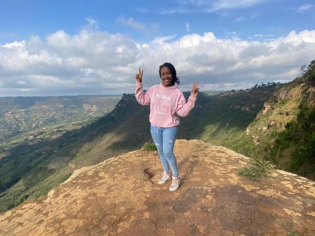 NaomiReyes stands on a rocky cliff, making double peace signs, wearing a pink hoodie, blue jeans, and white sneakers, with green hills and a blue sky in the background.