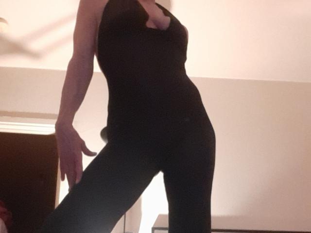 IssahneMay with a slim, toned physique, wearing a black, low-cut, sleeveless top and black pants, her small boobs visible, standing in a dimly lit room.