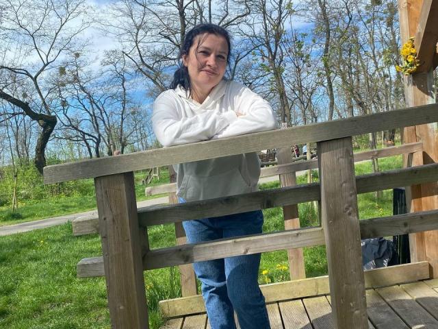 WestNancy leans on a wooden fence, wearing a white hoodie and blue jeans, with arms crossed, smiling slightly, standing in a grassy park on a sunny day.