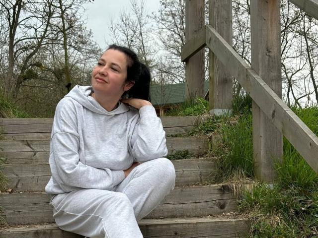WestNancy sits on wooden stairs in a gray hoodie and pants, hand in hair, smiling slightly with closed eyes, black hair, fair skin, and a relaxed, casual posture.