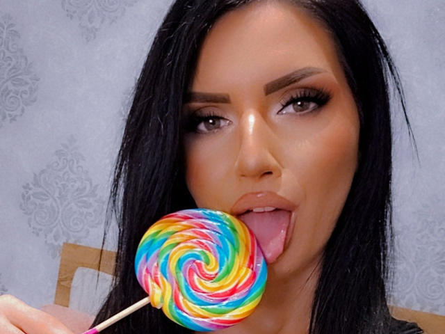 Paola Adams, con capelli neri, occhi marroni e trucco, con la lingua fuori, che lecca un lecca-lecca colorato, tenendolo con un bastoncino bianco nella mano destra..