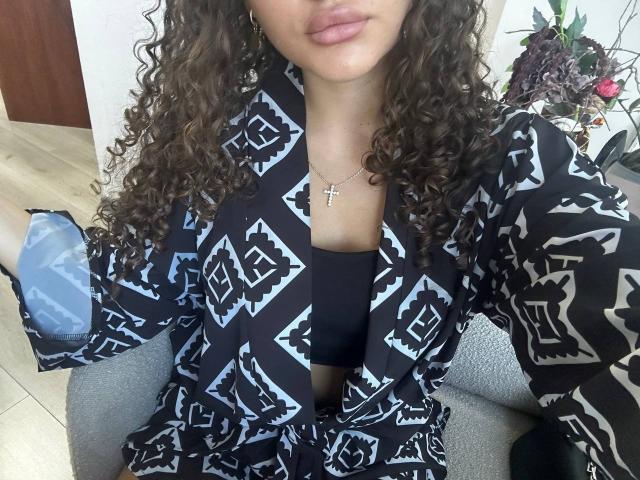 GloriaLi with curly brown hair, wearing a black patterned robe, black top, and cross necklace, showing her tits, holding a blue bag, sitting on a gray chair.
