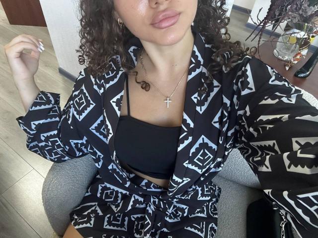 GloriaLi with curly brown hair, wearing a black geometric pattern robe, black bra, and gray leggings, showing her tits, sitting with a cross necklace, soft pink lips.