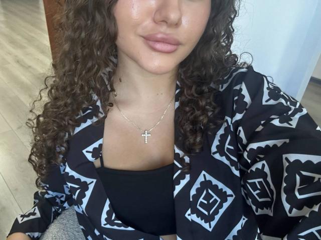 GloriaLi with curly brown hair, wearing a black and white patterned shirt, black bra, and silver cross necklace, pouting her pink lips, showing her subtle, shiny skin.