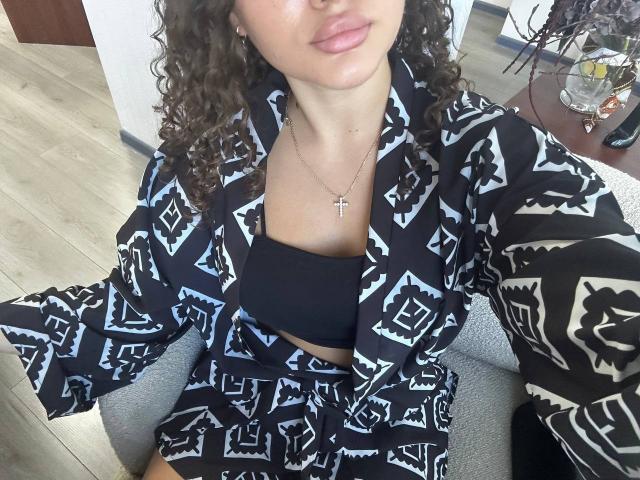 GloriaLi with curly brown hair, black and white geometric pattern robe, black tube top, matching robe and shorts, small cross necklace, sitting on gray chair, slightly parted lips.