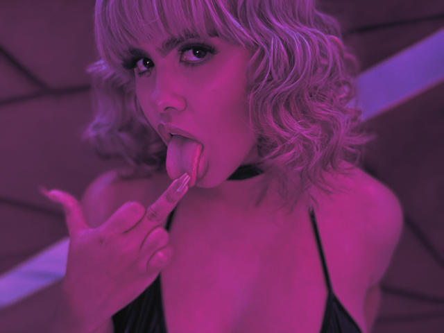 KhloeMartinez with pink-purple hair, tongue out, finger touching her lips, wearing a black choker and low-cut top, under pink lighting, showing her tits.