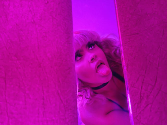 KhloeMartinez with blonde hair, choker, sticking out tongue, between two pink walls, purple lighting, looking up, visible breasts, sexy, playful, provocative, photo.