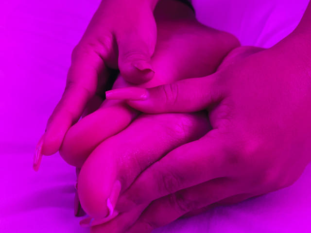 KhloeMartinez's hands with long pink nails gently hold her large, pink-lit boobs against a white background. Her fingers are slightly curled, showing soft skin and subtle texture.