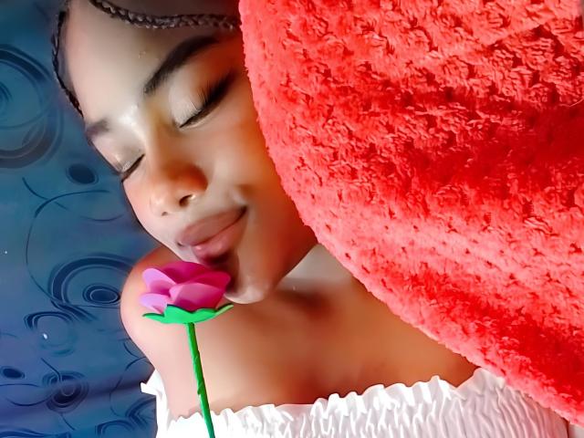 ShanissseRoss with eyes closed, smiling, holding pink rose to nose, red textured pillow on right, white off-shoulder top, blue background with swirls, brown braided headband.
