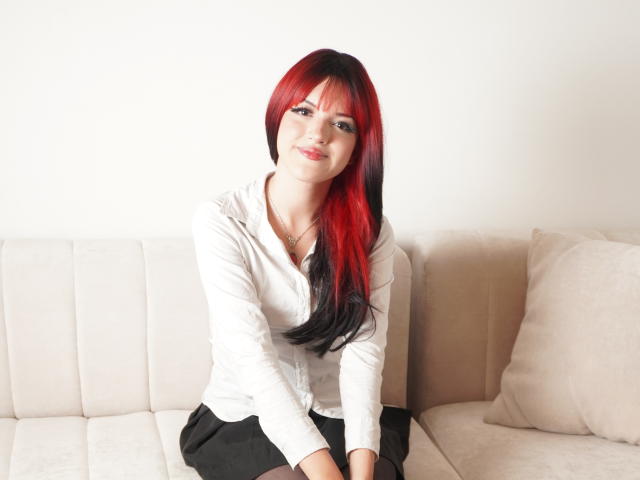 IrisWithmore has red and black hair, a white shirt, black skirt, and is sitting on a beige couch, smiling at the camera with a cute, sexy look.